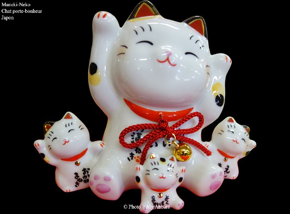 Maneki-Neko en famille, à fourrure blanche avec des taches noires et rousse ; ils lèvent soit la patte de devant droite, soit la gauche, soit les deux ; tous portent un collier rouge à petite clochette dorée