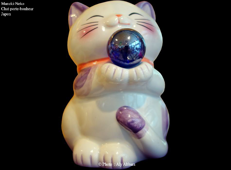 Une version commerciale moderne et simplifiée d'un Maneki-Neko composé d'un chat blanc avec du violet dispersé sur le corps ; un collier (écharpe) rouge autour du cou ; ce joyeux chat porte à l'aide de ses deux mains une grosse perle noire-violette